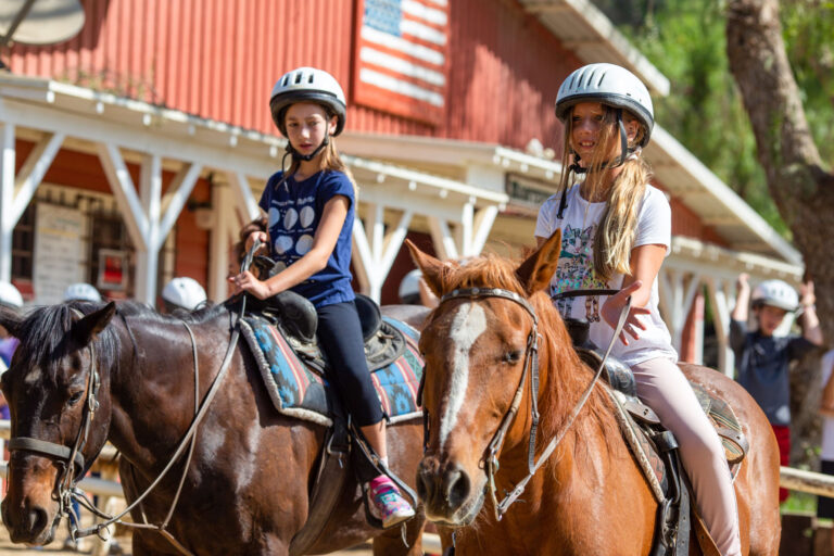 two girls riding horses in front of a red building.