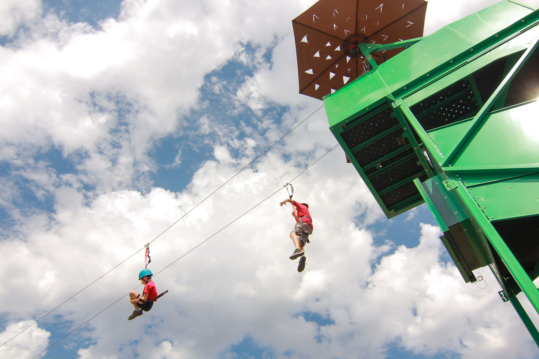 two kids riding down a zipline from a green tower.